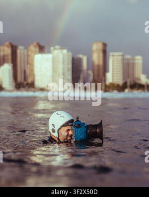 ALA MOANA BOWLS, HONOLULU, HAWAII - 10. APRIL 2022: Ein Unterwasserfotograf hält ein Gehäuse unter einem Regenbogen über der Skyline von Honolulu. Stockfoto