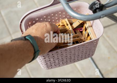 Nahaufnahme einer Hand, die eine Holzklammer aus einem hängenden Korb auswählt, der an einem Outdoor-Wäscheständer befestigt ist. In natürlichem Sonnenlicht aufgenommen Stockfoto