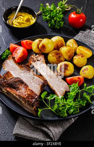 Amerikanische gegrillte Schweinerippchen aus dem Garten mit ganzen Kartoffeln, frischen Gartentomaten, krauser Petersilie und klassischem amerikanischem gelbem Ballpark-Senf auf dunklem Tee Stockfoto