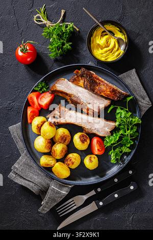 Bayrisch gebackene Schweinerippchen mit ganz gerösteten Kartoffeln, Tomaten, Petersilie auf einem Teller auf schwarzem strukturiertem Tisch mit Besteck und gelbem Senf,  ver Stockfoto