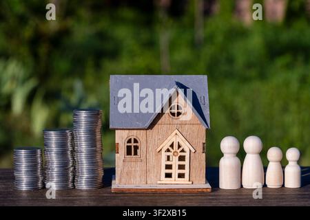 Holzhaus-Spielzeug zwischen Stapeln von Silbermünzen und symbolischer Holzfamilie von vier Personen auf dem Tisch platziert, Nahaufnahme. Immobilieninvestment, Hypothekenplanung Stockfoto