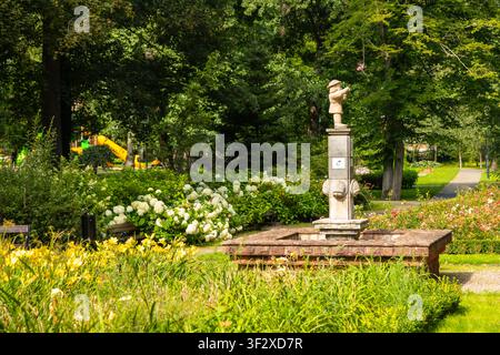 Ein steinerner Brunnen, der einen Jungen darstellt, steht in einem lebendigen Sommerpark, umgeben von Blumen, grünen Bäumen und einem fernen Kinderspielplatz, Polczyn-Zdro Stockfoto