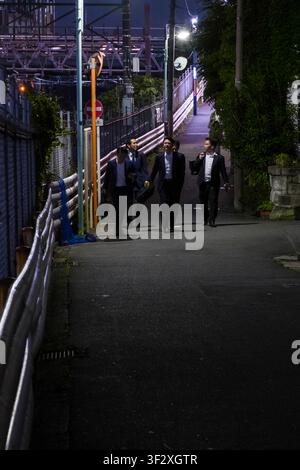 Diese Szene in Ebisuminami, Shibuya, Tokio zeigt eine Gruppe erwachsener Männer in Anzügen, die entlang einer gut beleuchteten Straße neben Bahngleisen gehen. Die Stockfoto