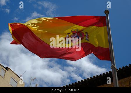 Spanische Nationalflagge mit Wappen, die vor einem blauen Himmel über einem Ziegeldach weht, Granada, Málaga, Andalusien, Spanien Stockfoto