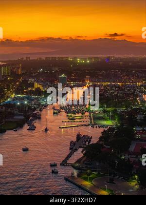 Luftbild der Marina Vallarta bei Sonnenuntergang mit zahlreichen Booten, die im Hafen vertäut sind, beleuchteten Stadthäusern und einem lebhaften orangefarbenen Himmel über dem Pazifik Stockfoto