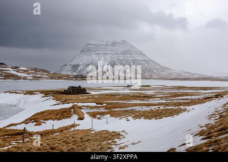Der majestätische schneebedeckte Berg Kirkjufell erhebt sich hinter einem zugefrorenen See in Island. Mitten in der goldenen Tundra mit Schnee steht ein kleines abgeno Stockfoto