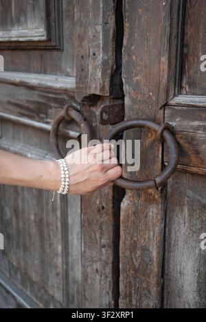 Frau öffnet verwitterte Holztür mit antiker Eisengriffschale, Fokus auf Hand und gealterter Textur. Erbe der Architektur, Geschichte, Handwerkskunst Stockfoto