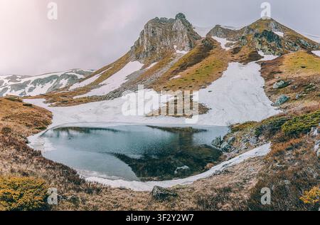 Beruhigende Berglandschaft mit einem teilweise gefrorenen Alpensee, der die umliegenden schneebedeckten Gipfel unter einem bewölkten Frühlingshimmel widerspiegelt Stockfoto