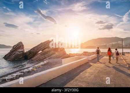 Touristen genießen einen Abendspaziergang an der Küstenpromenade, mit Sonnenliegen und auffälligen geschichteten Felsformationen am Strand Rafailovici in Montenegro Stockfoto