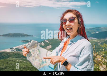 Glückliche junge Frau als Reisende, die eine Karte hält, auf Entdeckung zeigt und lächelt, während sie ein malerisches Sommerreiseabenteuer genießt Stockfoto