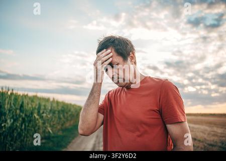 Mittelformat einer gestressten Person mittleren Alters, die auf einer ländlichen Schotterstraße neben einem Maisfeld steht und sich vor Schmerz die Hand an die Stirn hält. Selektiver Fokus. Stockfoto