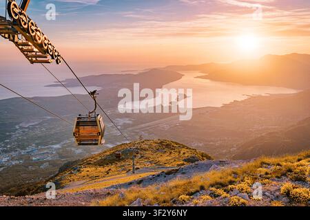 Seilbahn steigt einen schroffen Berghang hinauf, während ein dramatischer Sonnenuntergang die Bucht von Kotor und das Adriatische Meer unten beleuchtet Stockfoto