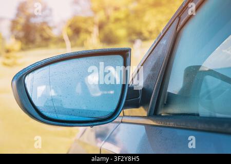 Makro-Nahaufnahme eines seitlichen Flügelspiegels eines dunklen Autos, bedeckt mit Regentropfen, der den Himmel mit verschwommenem grünen Blattwerk im Hintergrund reflektiert. Stockfoto