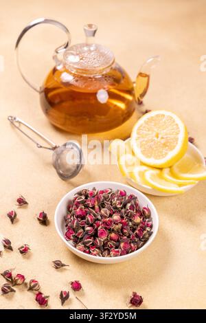 Glästeekanne mit Kräuter-Rosenknospen-Tee, begleitet von einer Schale getrockneter Rosenknospen, frischen Zitronenscheiben und einem Edelstahl-Teesieb auf einem hellen Ba Stockfoto