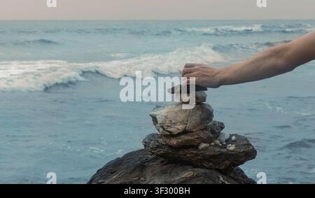 Eine Nahaufnahme eines menschlichen Arms und einer Hand, die sorgfältig einen Kieselstein auf einen Steinhaufen-Turm legen Stockfoto