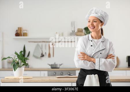 Lächelnde junge Köchin in weißer Uniform und Toque, die mit verschränkten Armen in einer modernen Küche steht, die professionelle Köchin strahlt Selbstvertrauen und Glück aus Stockfoto