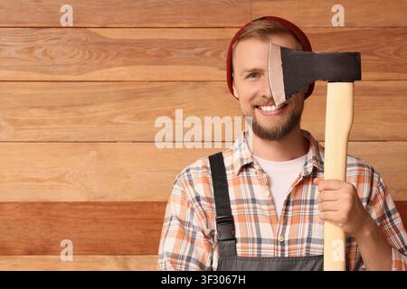 Lächelnder junger Mann in kariertem Hemd und roter Mütze, der eine Axt über seinem Auge gegen einen Holzhintergrund hält Stockfoto