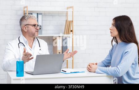 Serious Senior Mann in weißem Mantel und Brille spricht zu Patientin am Tisch Stockfoto