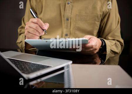 Mann schreibt mit der Hand auf einem Papier-Clipboard-Checklisten, während er mit einem Laptop auf dem Schreibtisch arbeitet. Konzept des Projektmanagements, des Ausfüllens von Umfragen, Businessplanni Stockfoto