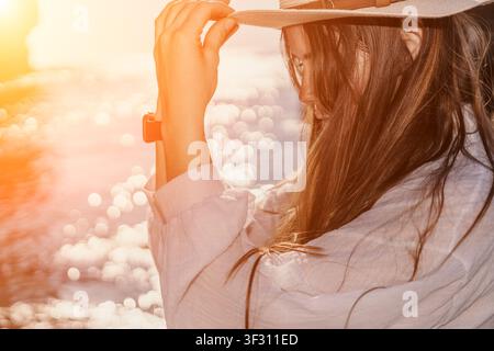 Frau mit Sonnenuntergangshut entspannt sich am Meer zur Goldenen Stunde in einem weißen Leinenhemd während eines friedlichen Sommerurlaubs Stockfoto