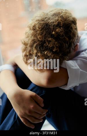 Vertikale Aufnahme eines kaukasischen Jungen, der mit dem Kopf auf den Knien sitzt, sein Gesicht verbirgt und sich in der Nähe eines Fensters zurückzieht, um Traurigkeit oder Stress auszudrücken. Nützlich für Kampagnen zu psychischer Gesundheit, Mobbing, Einsamkeit Stockfoto