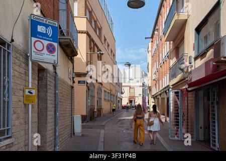 Viladecans, Spanien – 21. Juli 2026: Fußgänger gehen eine schmale städtische Gasse entlang, vorbei an einem Fotoüberwachungs-Straßenschild in Viladecans. Stockfoto
