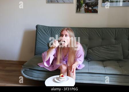 Frau isst Kuchen in einem gemütlichen Wohnzimmer, veranschaulicht Mäßigung, Selbstfürsorge und realistische Ernährungsgewohnheiten. Stockfoto