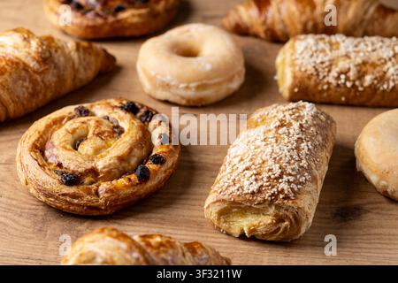 Auswahl an frisch gebackenen Gebäckstücken inklusive Croissants, Rosinenzwiebel, glasierten Donuts und süßen Gebäckstücken, die auf einem rustikalen Holztisch arrangiert sind. Stockfoto