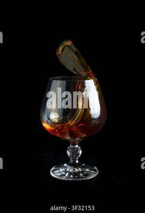 Ein dramatischer Spritzer von bernsteinfarbenem Cognac, der mitten im Gießen in einem Kristall-Brandy-Schnapsglas gegen einen tiefschwarzen Hintergrund eingefangen wird, und betont die Flüssigkeitsbewegung, Reflektion Stockfoto