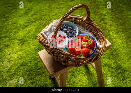 Korb aus Weidengeflecht, gefüllt mit frischen Erdbeeren, Himbeeren und Blaubeeren auf einem Holzstuhl Stockfoto