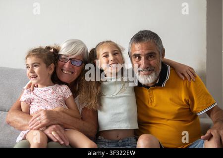 Zufriedene Großeltern und ihre Enkelinnen sitzen zusammen auf dem Sofa zu Hause und lächeln in die Kamera. Stockfoto