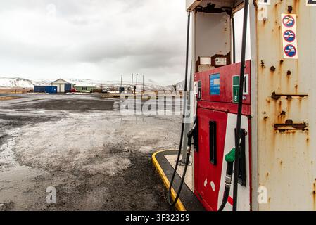 Nahaufnahme einer rostigen, vintage Kraftstoffpumpe im Vordergrund. Der Hintergrund zeigt einen verlassenen, nassen Asphaltplatz mit kleinen ländlichen Gebäuden und Schnee Stockfoto