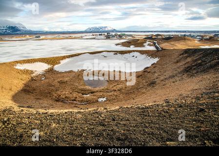 Die Ansicht vom Rand zeigt das Innere eines Vulkankraters mit trockenem Gras, einer Pfütze und Schneeflecken. Im Hintergrund erstreckt sich ein gefrorener See, Stockfoto