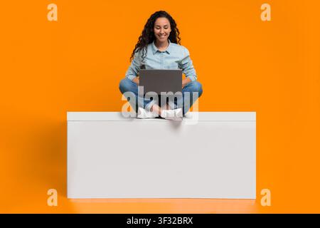 Frau sitzt auf weißem Kasten und nutzt einen Laptop vor einem hellen orangefarbenen Hintergrund am Tag Stockfoto