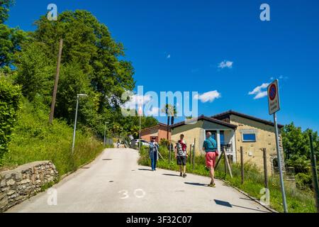 Caslano – Schweiz – 12. Mai 2026: Touristen gehen entlang einer Dorfstraße an einem Verkehrsschild, einem Steinbau und Grünflächen vorbei, Malcantone, Tessin. Stockfoto