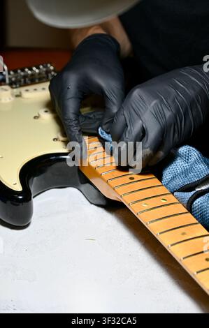 Luthier-Hände in Nahaufnahme eines Gitarren Technikers oder Luthiers, der schwarze Nitrilhandschuhe trägt, während er einen E-Gitarren-Ahornhals und Griffbrett reinigt, poliert und einstellt. Makrodetail der Gitarrenwartung, Fretarbeit, Luthierwerkstatt und Instrumentenpflegeprozess. Ideal für Musikdienstleistungen, Reparaturwerkstatt, Rockmusik und Handwerkskonzepte. Schutzhandschuhe reparieren und polieren das Griffbrett der E-Gitarre. Hochwertiges Foto Stockfoto