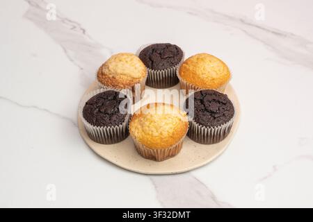 Sechs Vanille- und Schokoladen-Muffins, die in einem Kreis auf einem gesprenkelten Sahneteller über Marmor angeordnet sind. Stockfoto