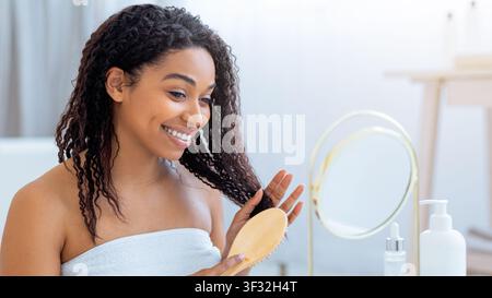 Lächelnde junge afroamerikanische Frau, die sich zu Hause vor dem Spiegel mit einer Bürste um ihr lockiges Haar kümmert Stockfoto