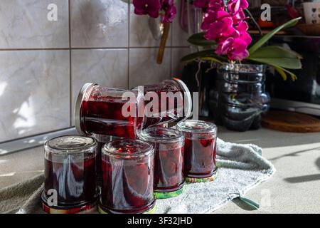 Gemütliche Küchenkomposition mit dunkelroter Kirschmarmelade in Gläsern, frischen Blumen und rustikaler Einrichtung für hausgemachte süße Beerenkonfitüre Stockfoto