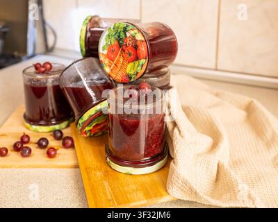 Rote Stachelbeer-Marmelade im Glas mit Metall Deckel auf weißem Stoffserviette, gemütliche rustikale Küchenausstattung mit frischen Beeren, hausgemachte Konserven Stockfoto