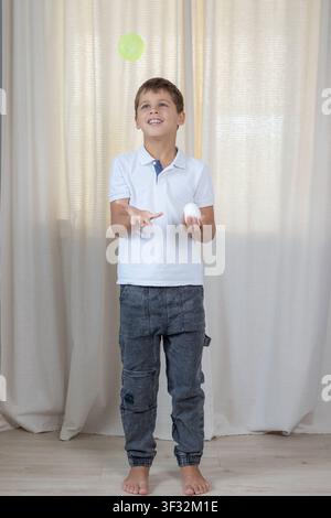 Lächelnder Schuljunge jongliert drinnen mit grünem Ball, einen weißen Ball in der Hand haltend, trägt weißes Polohemd Stockfoto