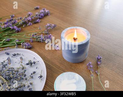Duftender Zementkerzenbrenner auf Holztablett in der Nähe eines Lavendelstraußes. Keramikplatte mit getrockneten Blumen im Hintergrund liegend. Gemütliches Aromatherapie-Spa Stockfoto