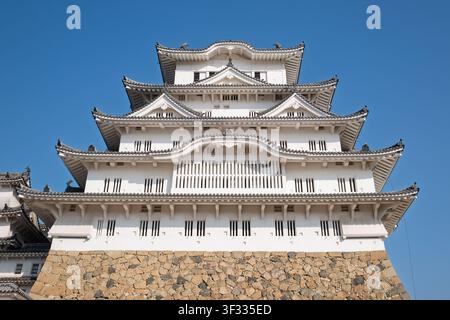 Direkte Frontansicht aus niedriger Perspektive der symmetrischen, gestuften weißen Fassade und der goldenen Turmspitzen des Hauptgebäudes des Himeji-Schlosses, Japan. Stockfoto