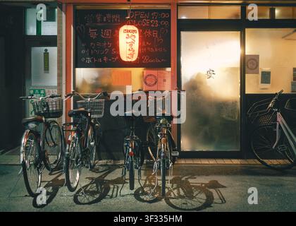 Lebendige Nachtansicht eines kleinen lokalen Izakaya-Restaurants mit einer beleuchteten rot leuchtenden Papierlaterne und geparkten Fahrrädern, Osaka, Japan Stockfoto