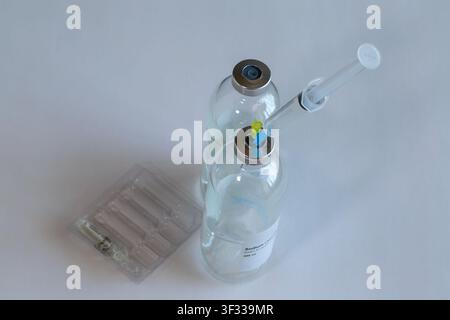 Zwei klare Glas-Medicinfläschchen mit Kochsalzlösung für IV-Tropfinfusionen auf einem weißen Tisch, mit einer eingeführten Spritze und Ersatznadeln in der Nähe. Stockfoto