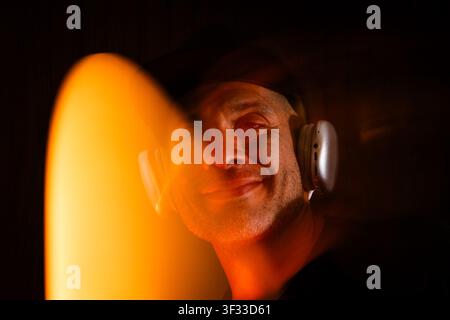 Mann lächelt, hört Musik über Kopfhörer, umgeben von orangefarbenem Licht und Unschärfe Stockfoto