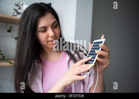 Junge Frau verwendet Smartphone mit tragbarem Solarladegerät im Innenraum. Technologie der erneuerbaren Energien und nachhaltige mobile Stromlösungen. Stockfoto