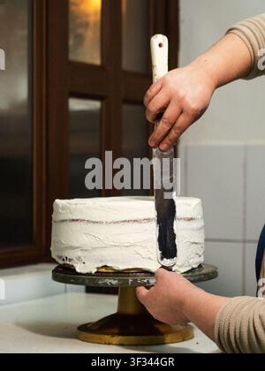 Hände glätten weißen Zuckerguss auf einem runden Kuchen. Ein Keramik-Kuchenständer hält das Dessert. Ein goldener Sockel glänzt im warmen Licht. Eine Person streicht vorsichtig die Glasur Stockfoto