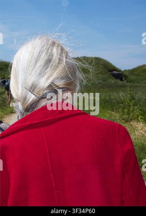 Rückansicht einer Frau mit grauen Haaren, die einen hellroten Mantel trägt, während sie durch eine grasbewachsene Küstenlandschaft geht Stockfoto