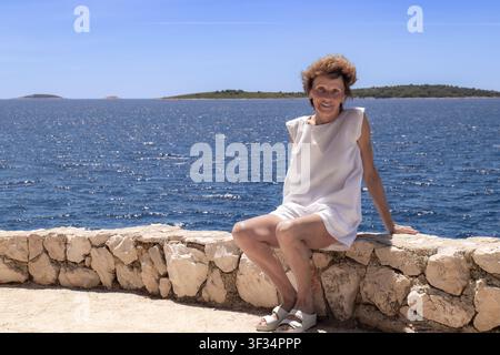 Zufriedene Seniorin in weißem Sommerkleid sitzt auf einer Steinmauer an der Adria, Kroatien Stockfoto